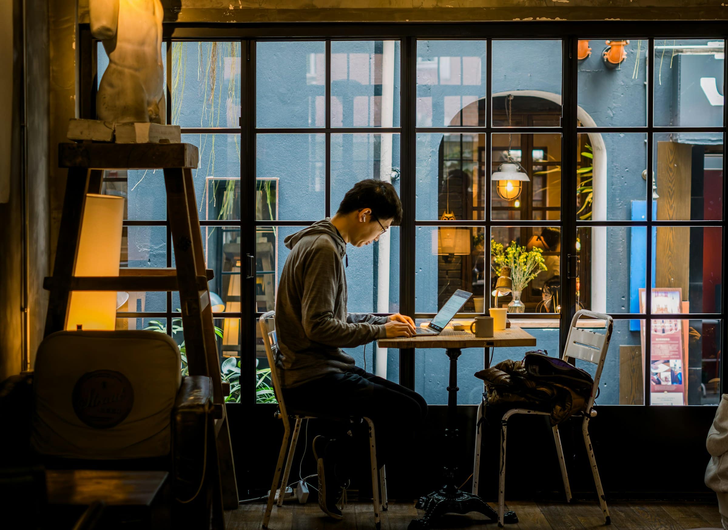 A designer working on a laptop at a warmly lit table beside a large window on a cold blue evening